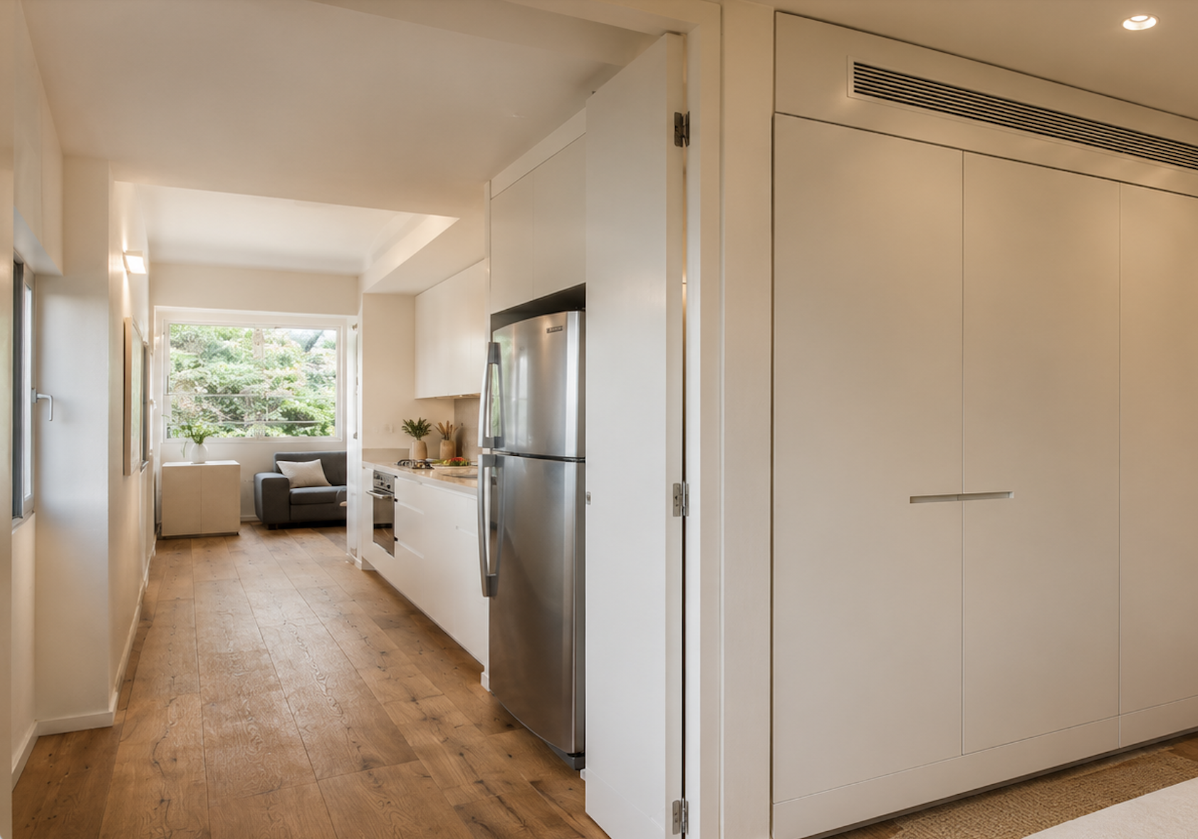 Bright modern apartment hallway with built-in white storage, stainless steel refrigerator, wood flooring and living area beyond